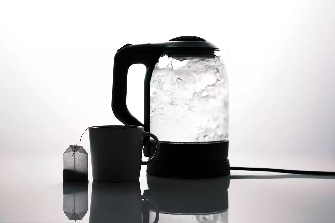 A minimalist setup featuring a glass electric kettle boiling water next to a simple white mug with a tea bag placed beside it. The scene is set against a bright, white background.