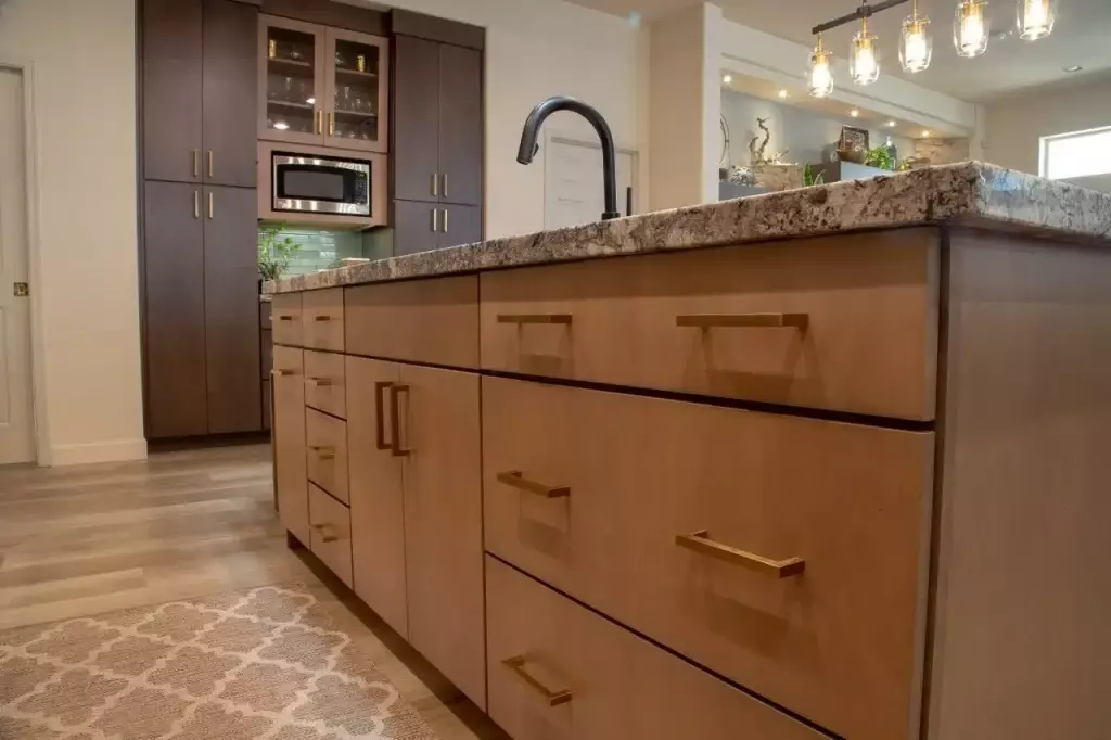 Close-up view of a modern kitchen island with sleek natural wood drawers, brushed gold hardware, and a granite countertop, complemented by dark wood cabinetry and under-cabinet lighting in the background.