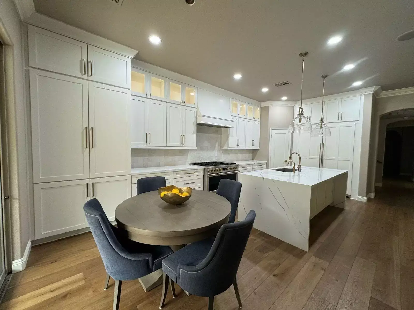 Modern kitchen interior featuring slip-resistant wood-look flooring, white cabinetry, a marble island, and a round dining table with navy upholstered chairs.