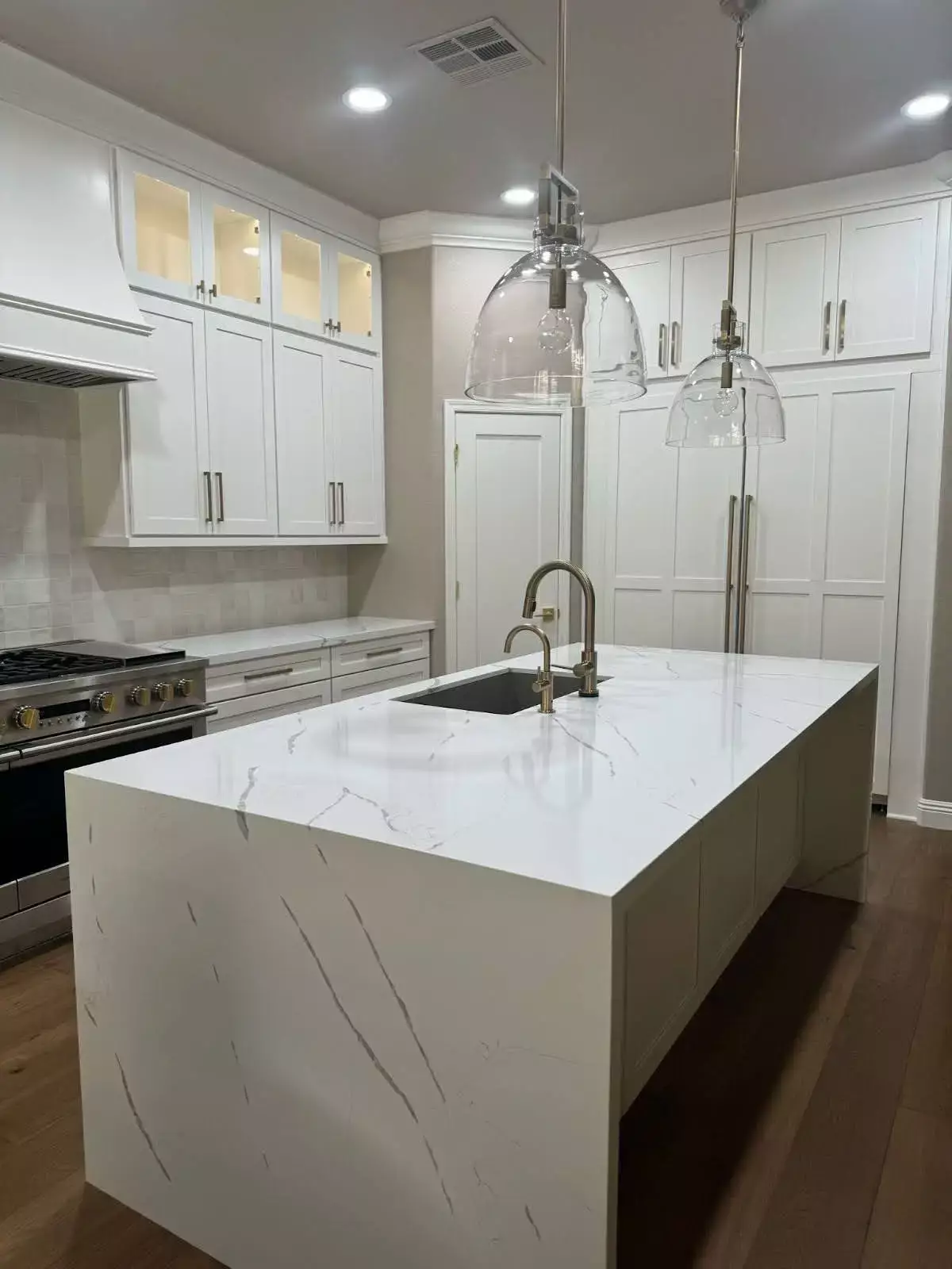 Elegant white kitchen with waterfall marble island, integrated sink, glass pendant lights, gold hardware, and seamless built-in cabinetry—blending appliances into a sleek, contemporary design.